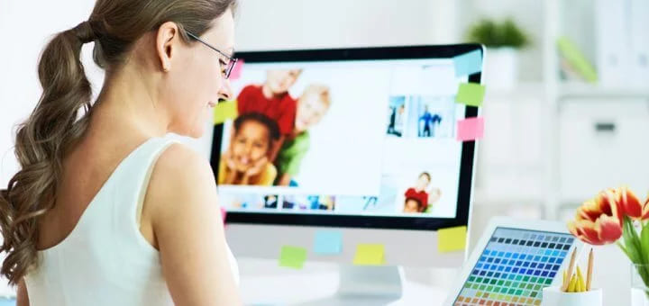 A woman at a desk reviewing photos on a monitor framed by sticky notes, with a colour swatch chart beside her