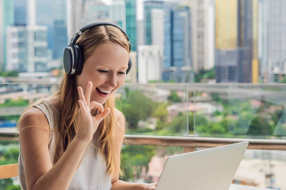 A woman in headphones on a balcony making an OK gesture at her laptop, with a city skyline behind her