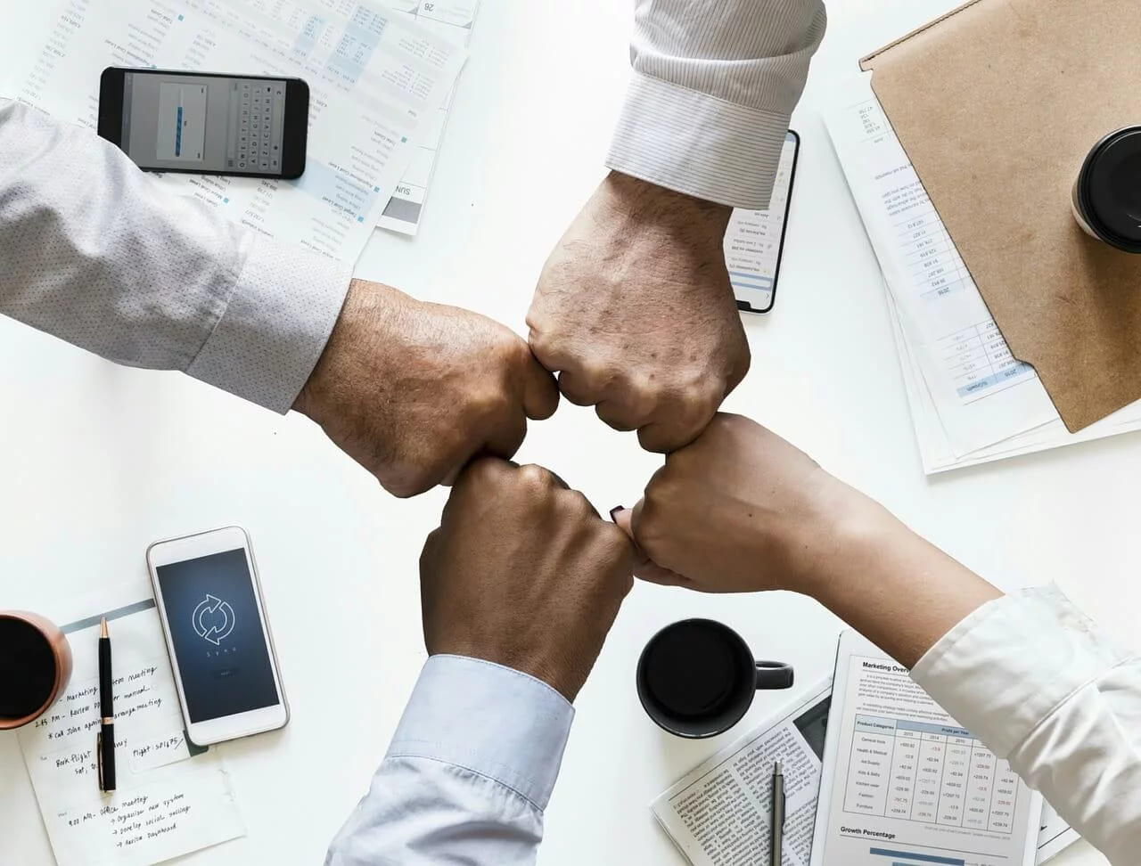 Four hands bumping fists over a desk covered with phones, coffee cups and papers