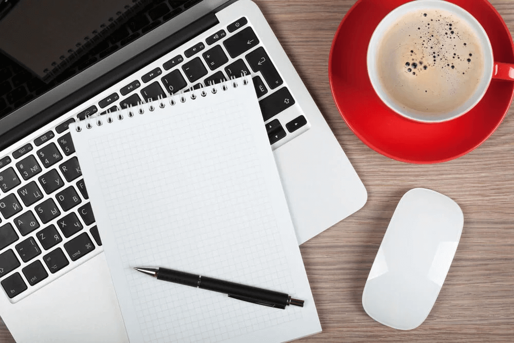 A blank notepad and pen on a desk beside a laptop, a mouse and a red cup of coffee