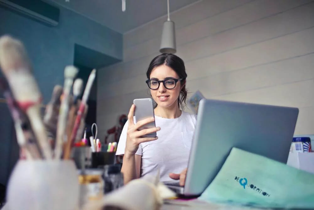 A woman in glasses holding her phone at a workshop desk, with a laptop and jars of paintbrushes around her