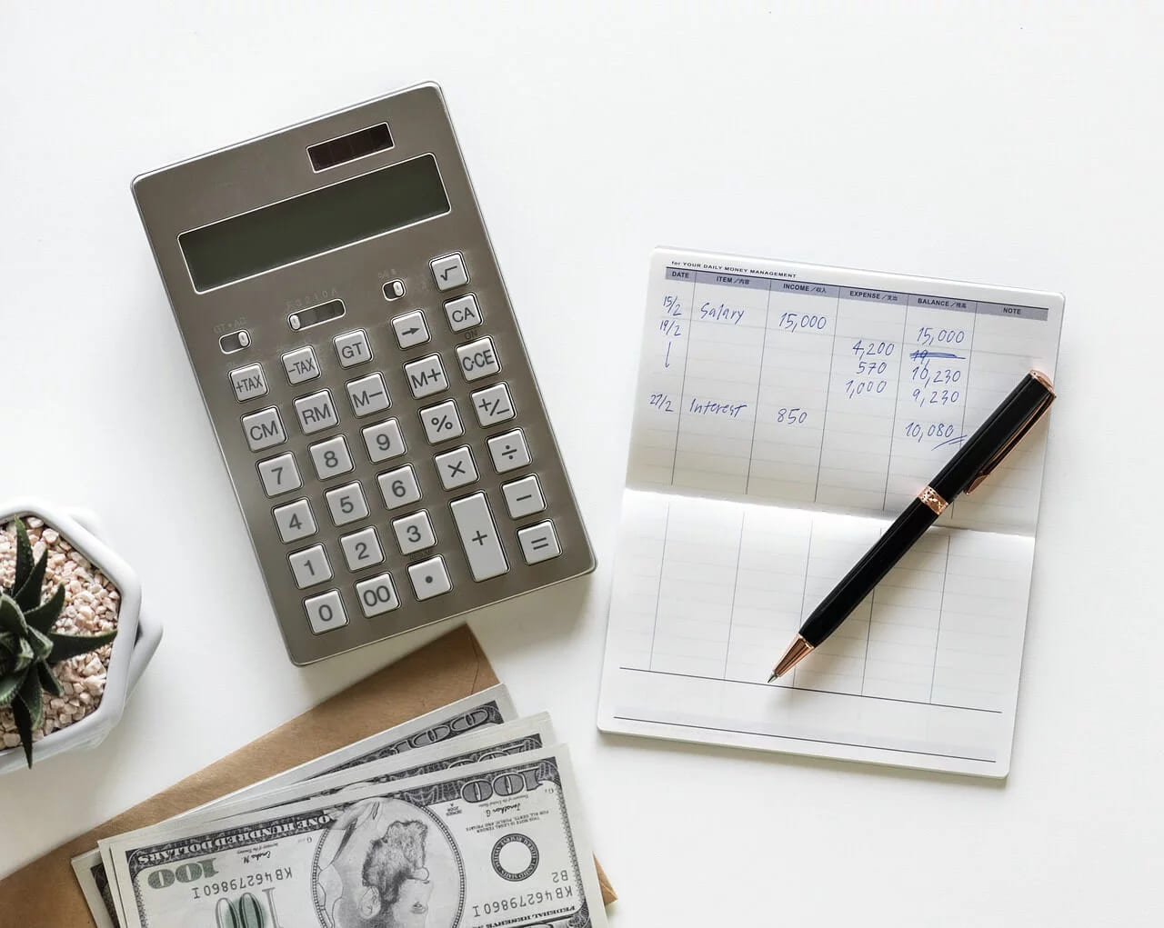 A calculator, banknotes and a pen beside a notebook of handwritten figures