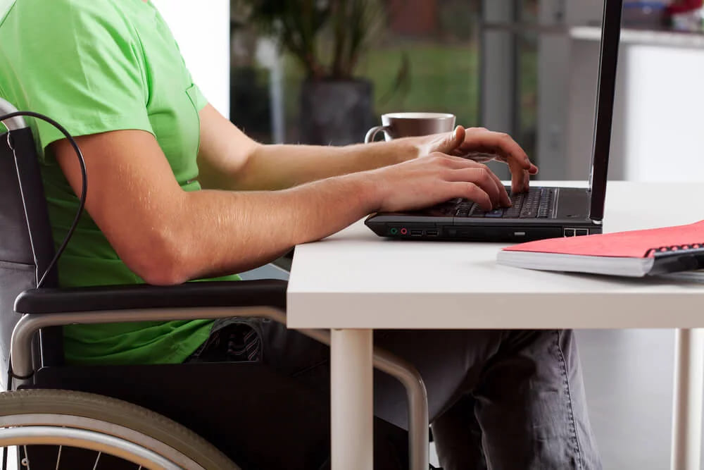 A person in a wheelchair typing on a laptop at a desk with a cup and a notebook