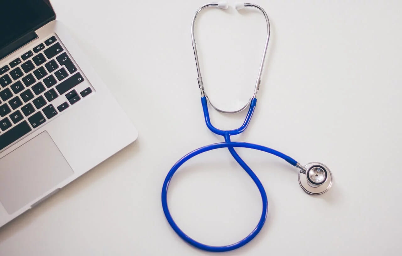 A blue stethoscope lying beside an open laptop on a white desk