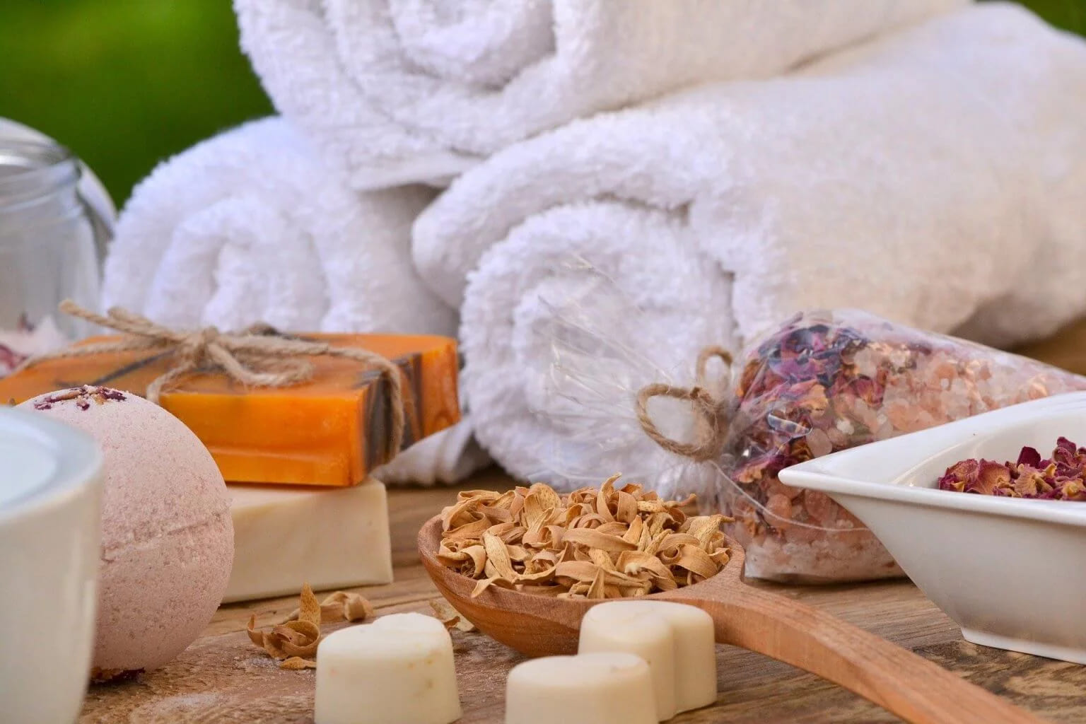 Home spa ingredients on a table, with rolled towels, a bath bomb, handmade soap, dried petals and bath salt