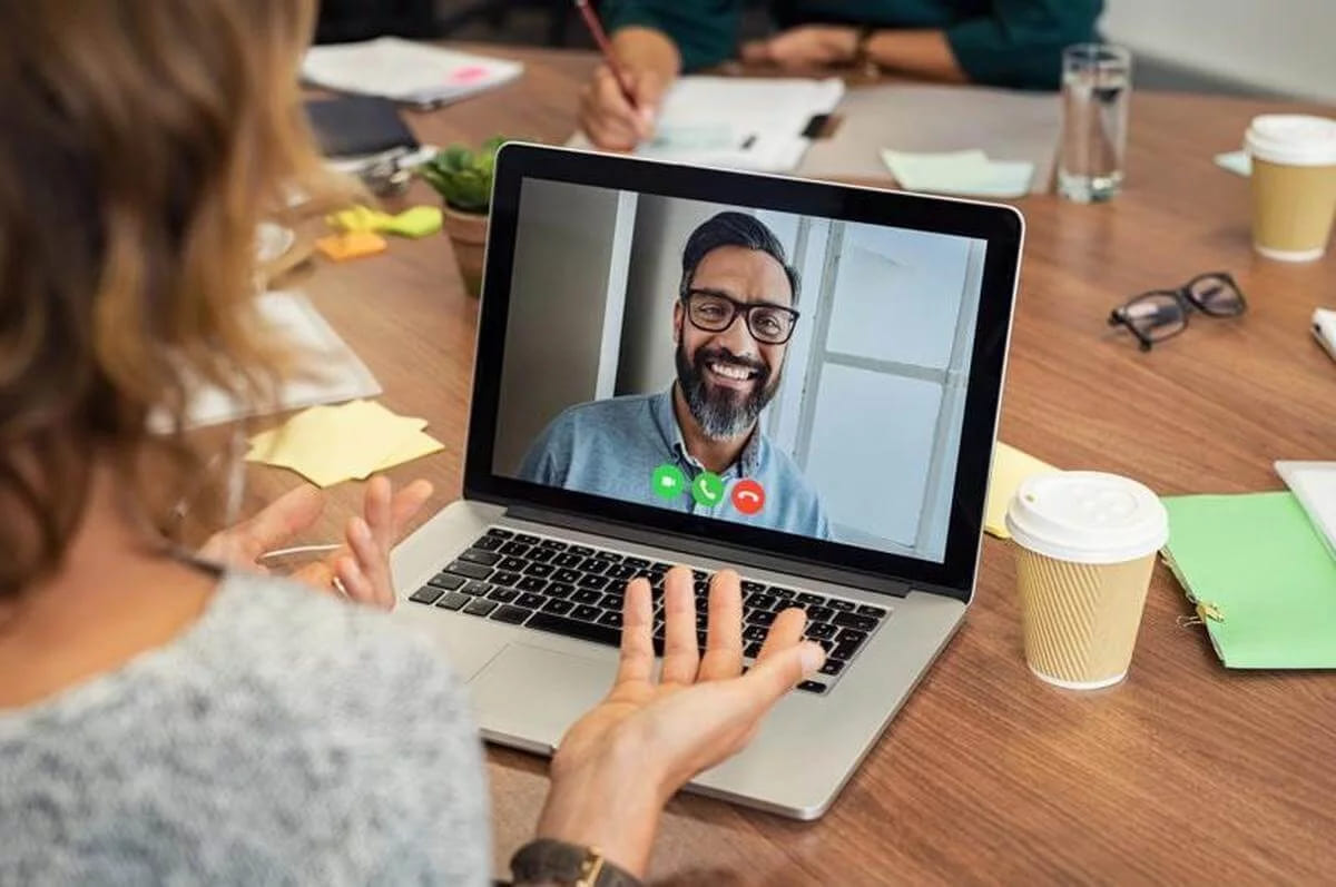 A woman at a laptop in an office waving at a smiling man on a video call, with the call buttons under his window and sticky notes and coffee cups on the table