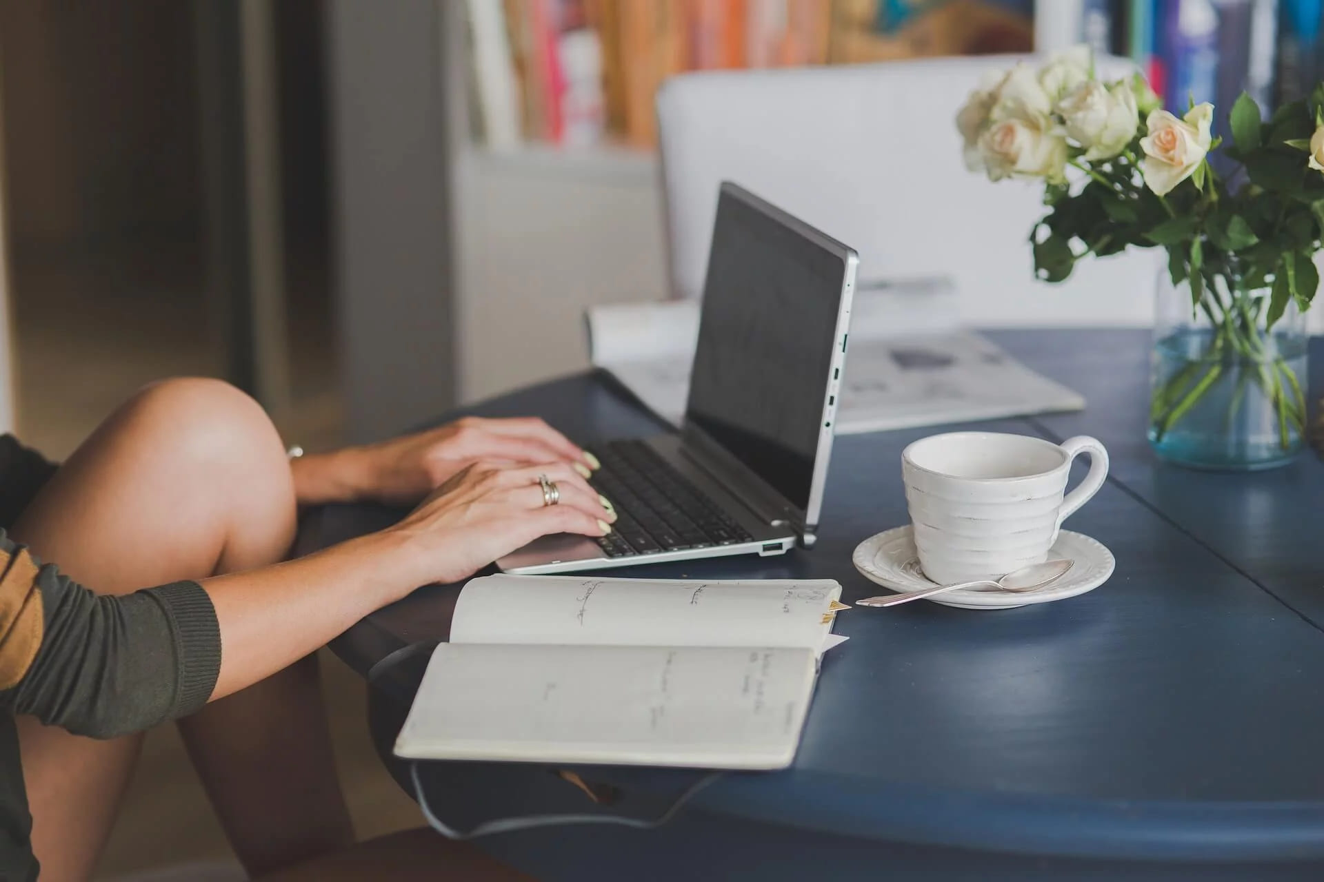 A woman typing on a laptop at a table with an open notebook, a cup and flowers