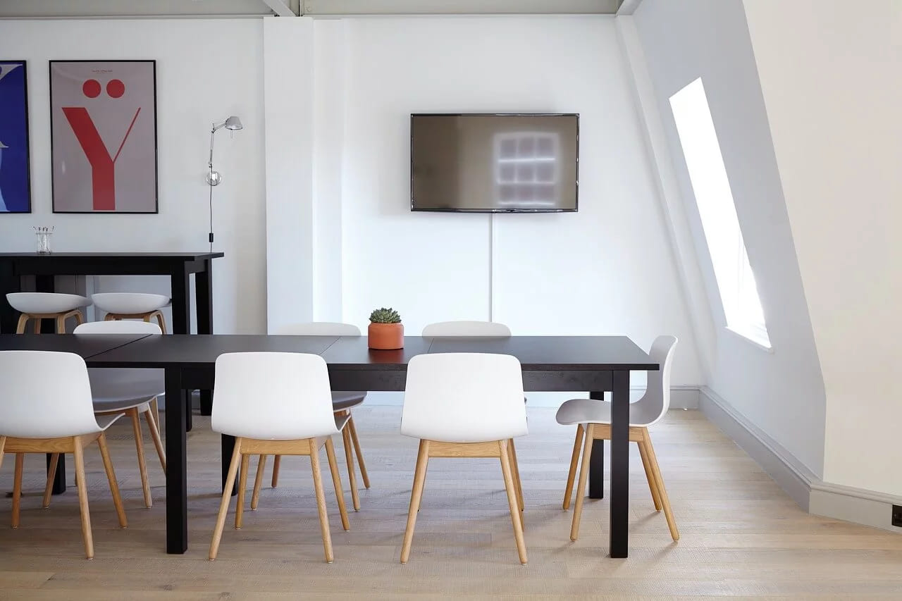 An empty meeting room with a long dark table, white chairs and a screen mounted on the wall