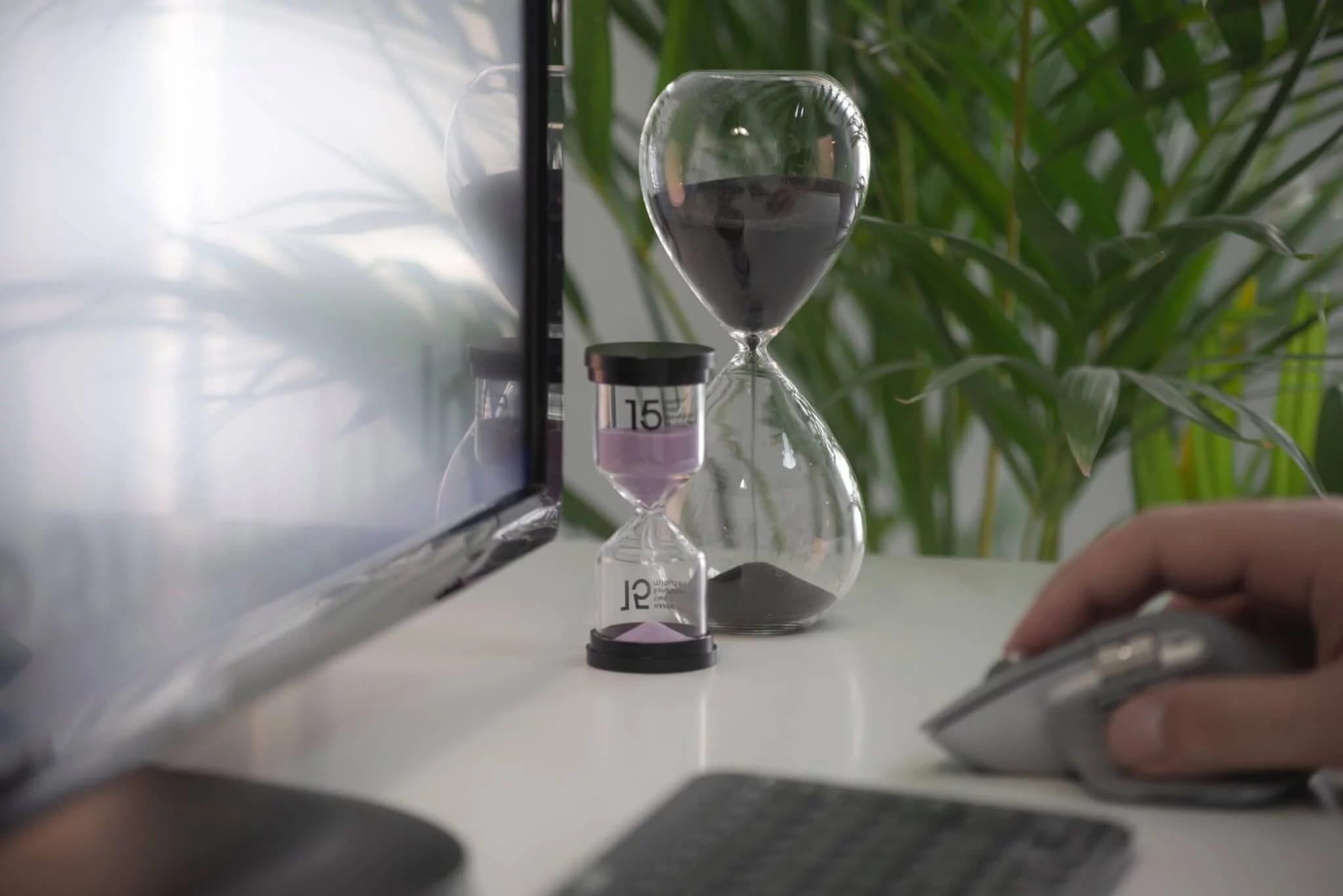 An hourglass standing on a desk beside a monitor while a hand rests on the mouse