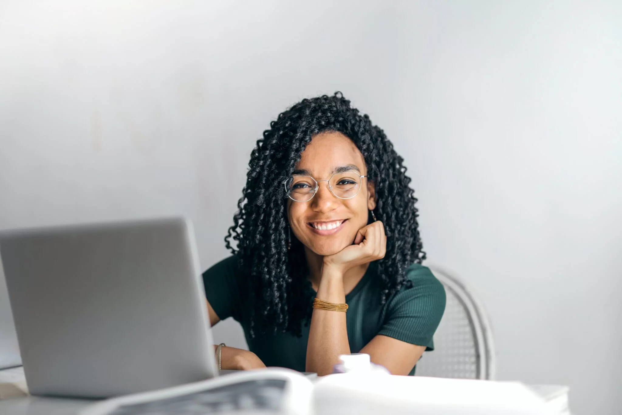 A smiling woman in glasses resting her chin on her hand at a desk with a laptop and an open book