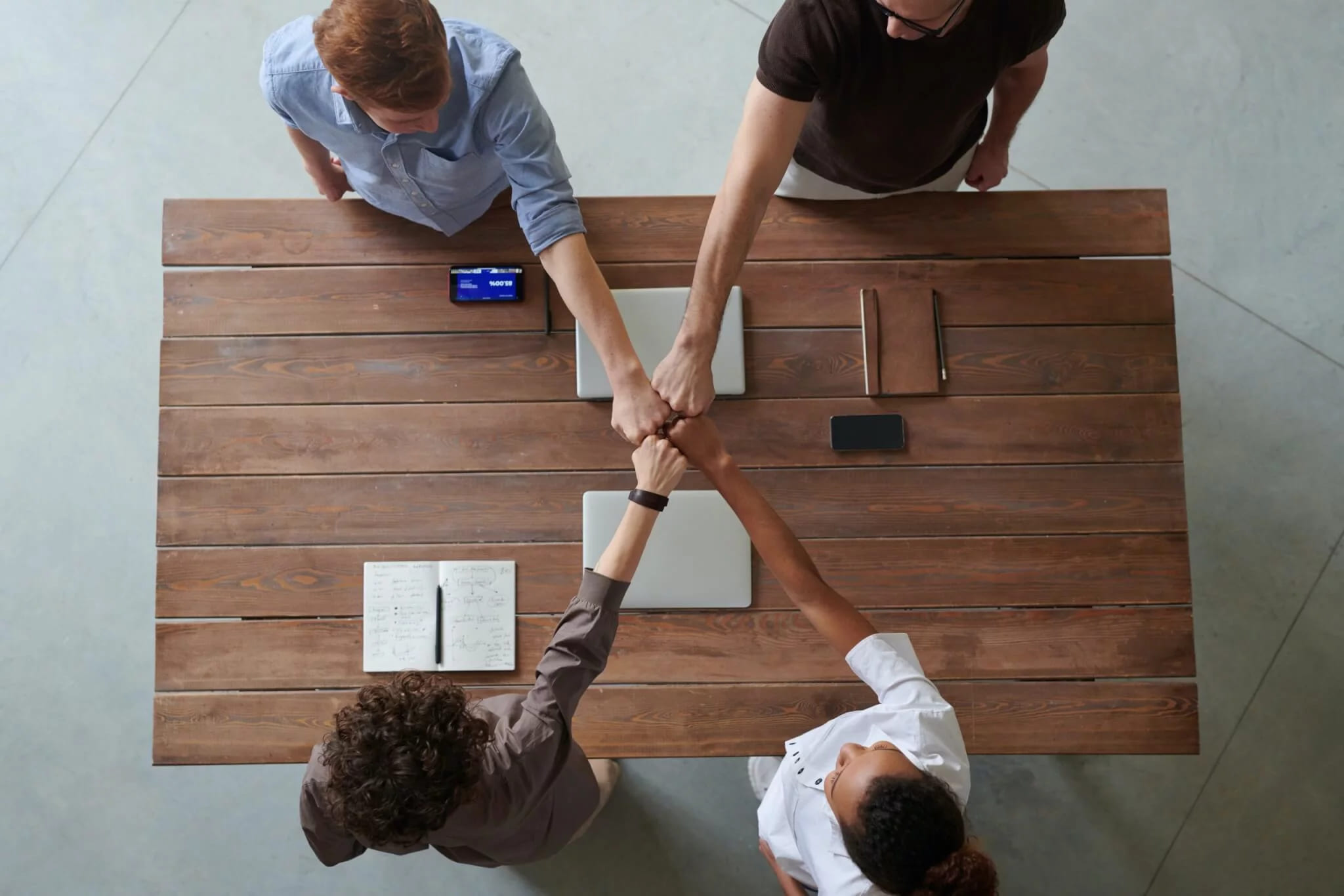 Four people around a wooden table seen from above, bumping fists in the middle, with laptops, phones and an open notebook beside them