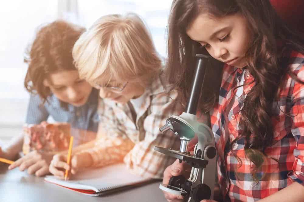 Three schoolchildren at a science lesson, one adjusting a microscope while the others write in a notebook