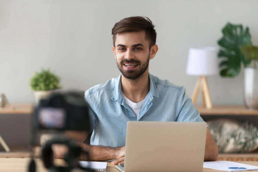 A man in a plain blue shirt at his laptop being filmed by a camera on a tripod
