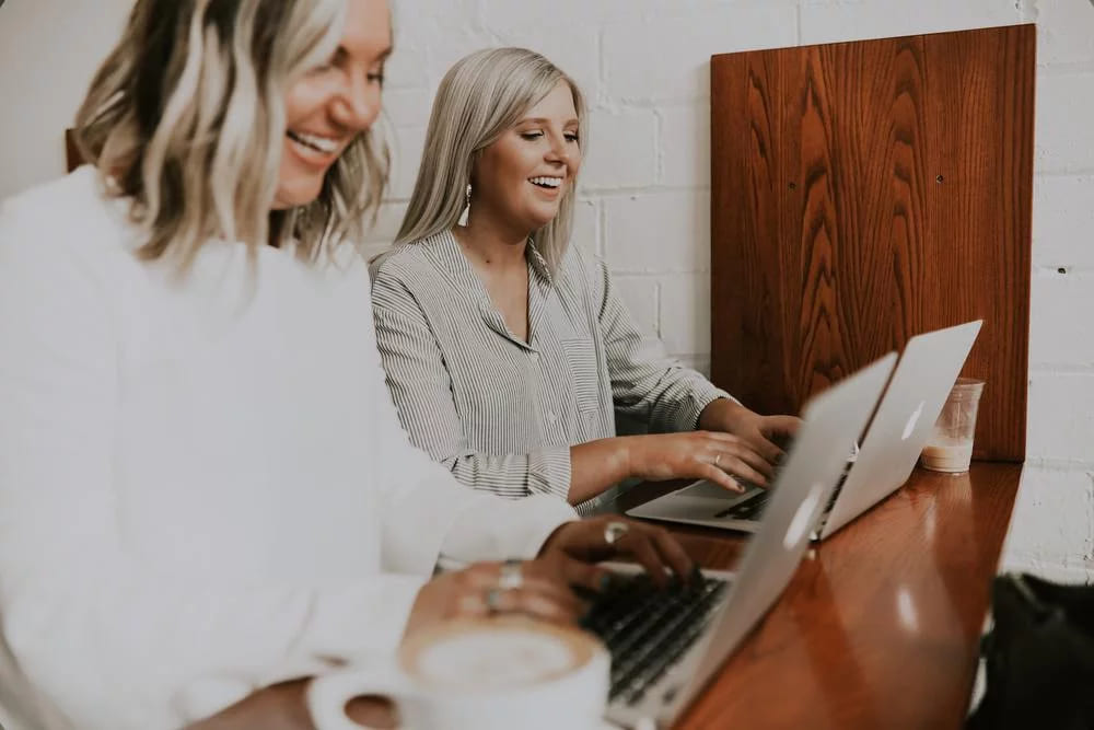 Two women laughing together at a laptop over coffee