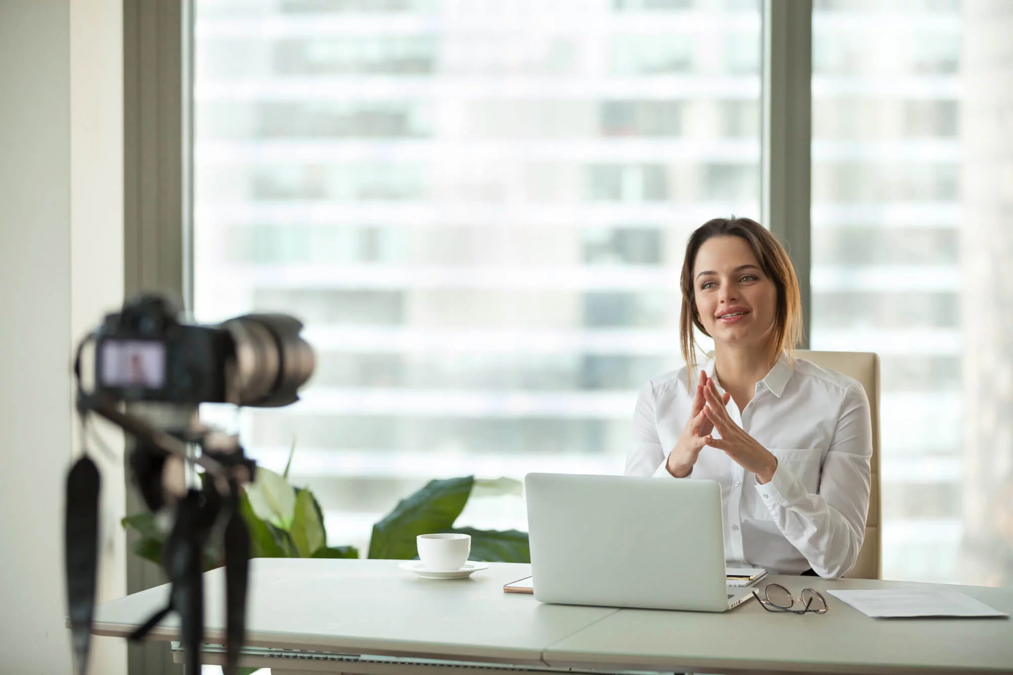 A woman at a desk with a laptop being filmed by a camera on a tripod