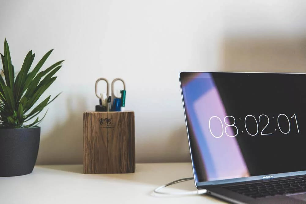 A laptop on a desk showing a large clock, beside a wooden pen holder and a potted plant