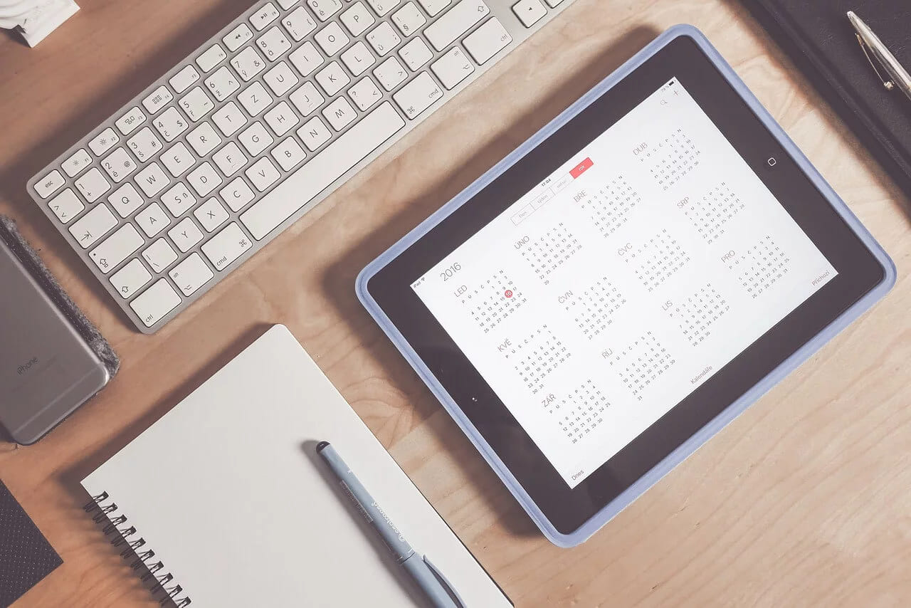 A tablet showing a monthly calendar beside a keyboard, a notebook and a pen