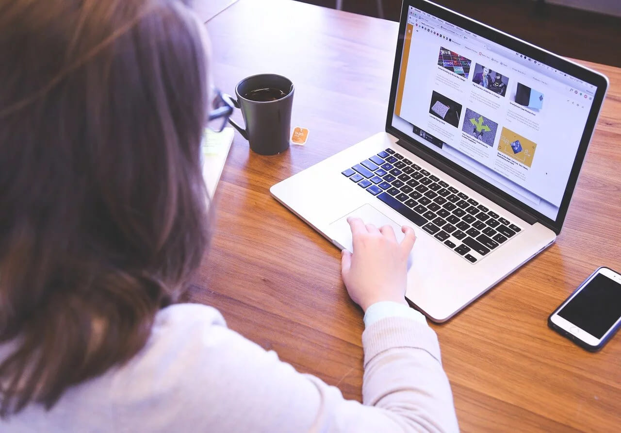 A woman at a laptop with a mug and a phone on a wooden desk