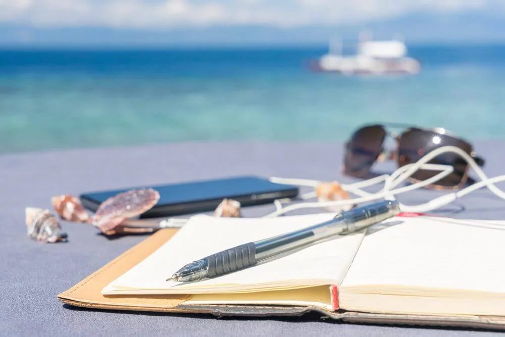 An open notebook, a pen, a phone and sunglasses on a table by the sea