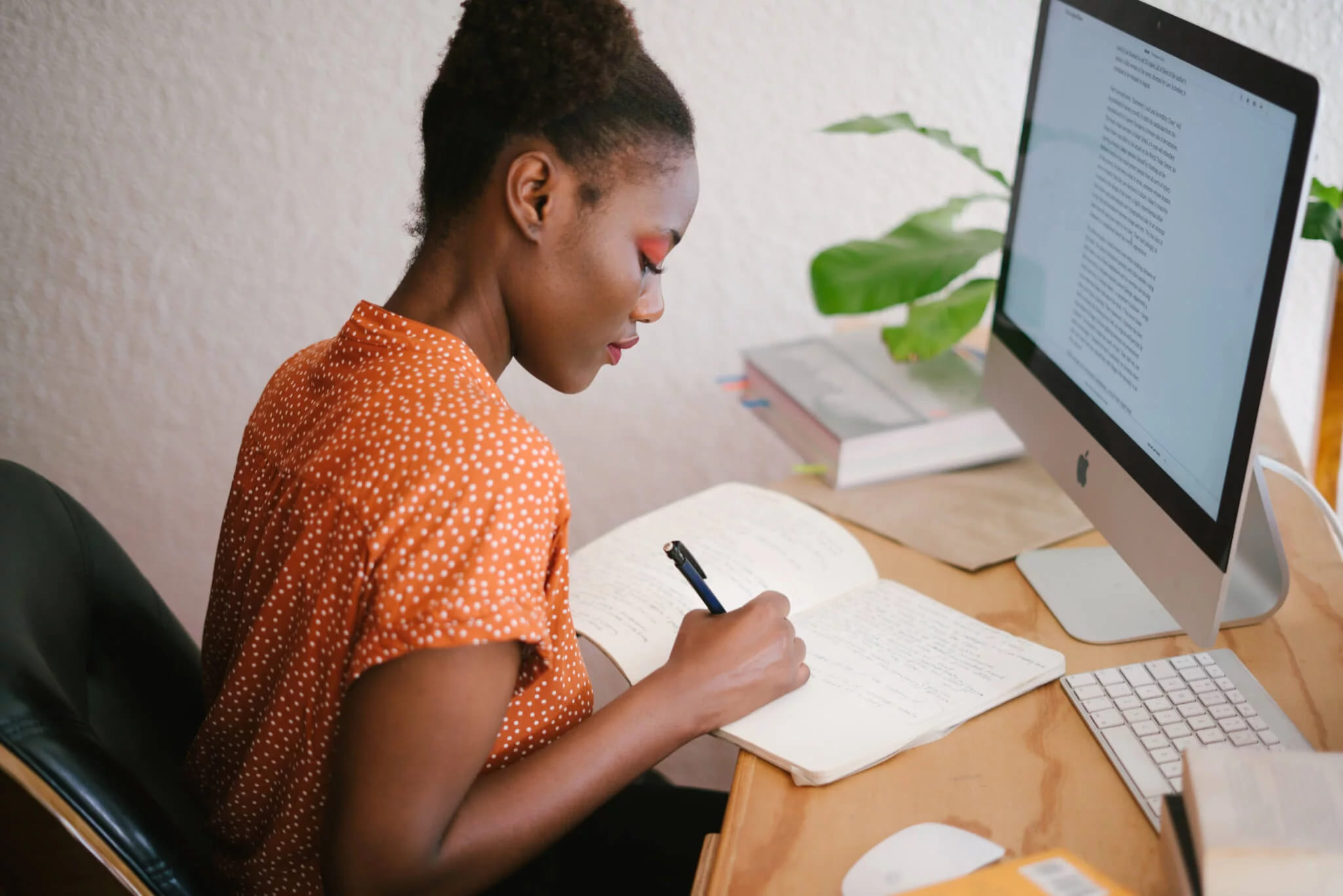 A woman writing in a notebook at a desk with a large monitor