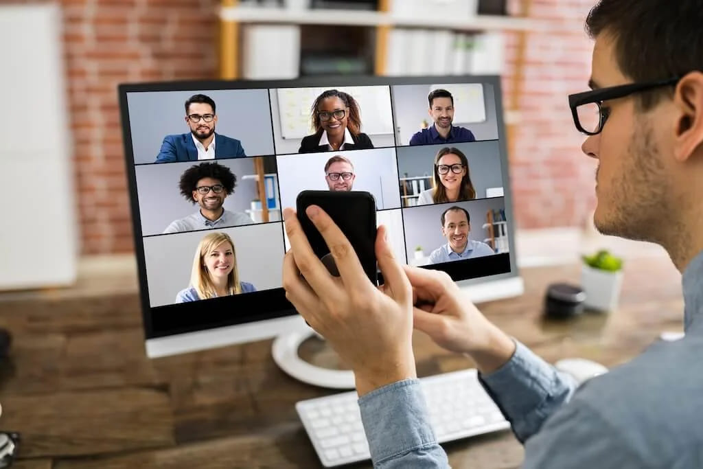 A man at a monitor showing a nine-person video call while holding his phone