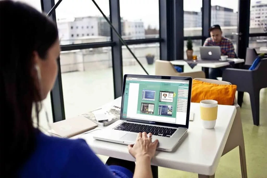 A woman working on a laptop at a white table by a window, with another person at a laptop in the background